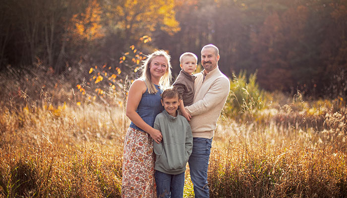 A mother, a father carrying a baby, and their eldest son stand in a field as the sun shines.