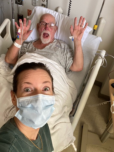 A person wearing a face mask in a hospital takes a selfie with a patient who has both of his hands raised with a funny face while he is on a hospital bed in the background