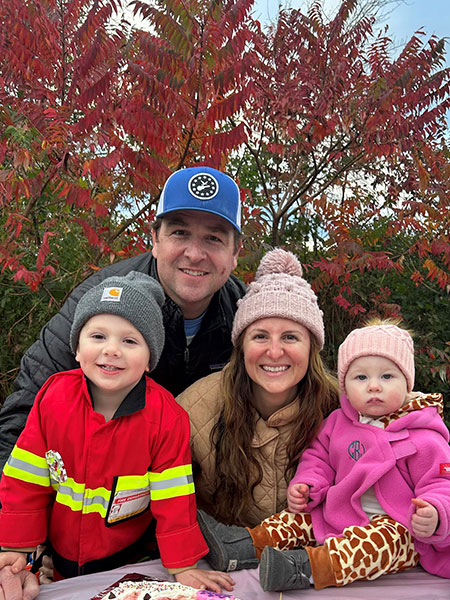 A photo of a family of four. consisting of a mother, father, and their two kids, taken during fall season with trees in the background